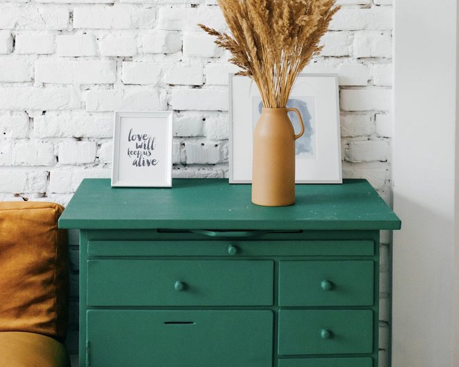 A green dresser with two white frames, one written 'love will keep us alive' and other with a painting that is not beeing completely visible because of a brown pot with hay in front of it